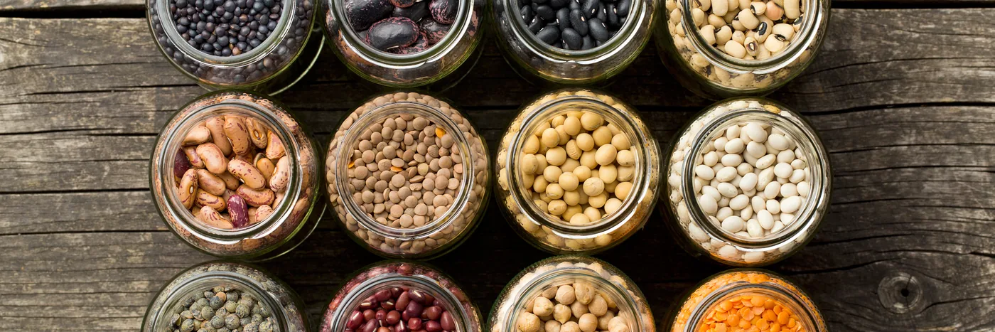 Glass jars of dried pulses, grains and seeds lined up in rows on a dark wooden shelf.